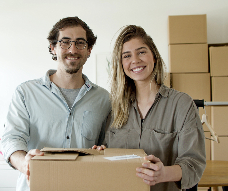 A smiling couple holding a cardboard box in a room filled with similar boxes, representing their thriving Amazon business. Their confident and relaxed demeanor suggests the ease with which they manage their online enterprise, thanks to the services provided by Prep Ninjas FBA Prep Center.