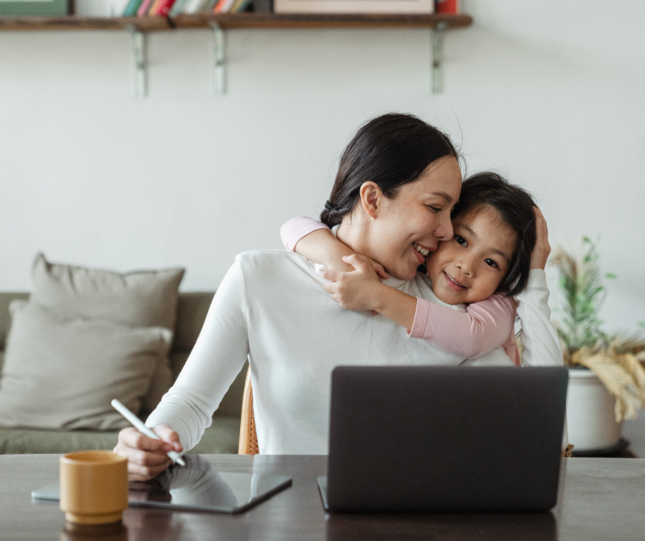 A woman working from home on her laptop with a digital tablet and stylus beside her, sharing a tender moment with her young daughter who is hugging her from behind. They are both smiling, embodying a warm, work-life balance as the woman manages her Amazon FBA business because of using Prep Ninjas as her FBA Prep Center.