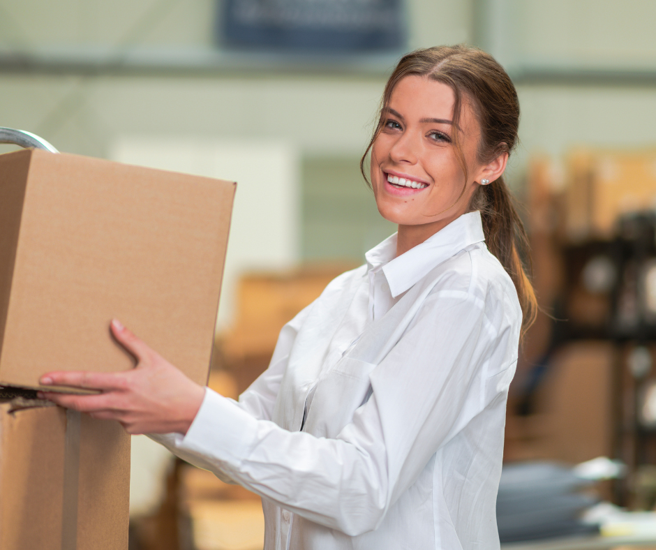 A cheerful woman with her hair tied back is standing in a warehouse, holding a cardboard box. She is wearing a white lab coat, suggesting she may be a worker or a supervisor. The background is slightly blurred, emphasizing her smiling face as the focal point.